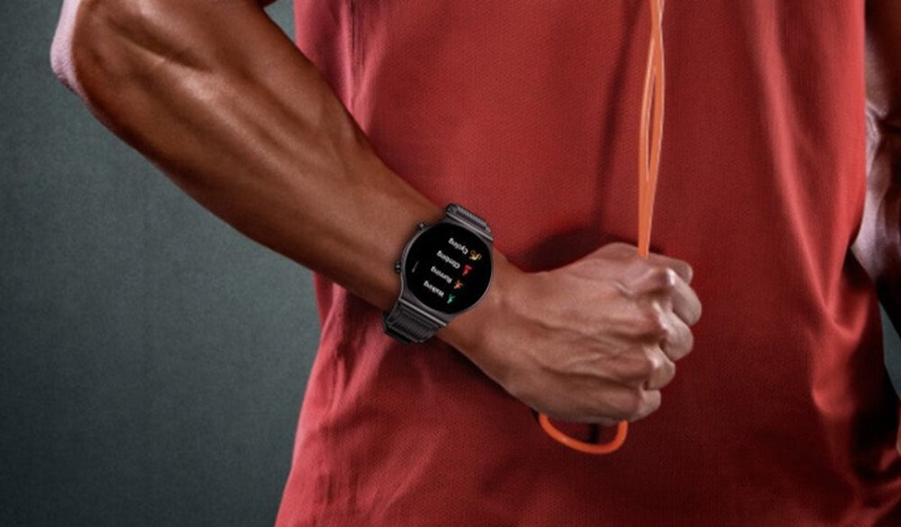 Osoba w czerwonej koszulce trzyma pomarańczową skakankę, na nadgarstku ma czarny smartwatch z wyświetlaczem pokazującym tekst. Na ekranie widoczne są opcje: \'Sunday\', \'Running\', \'Biking\'.