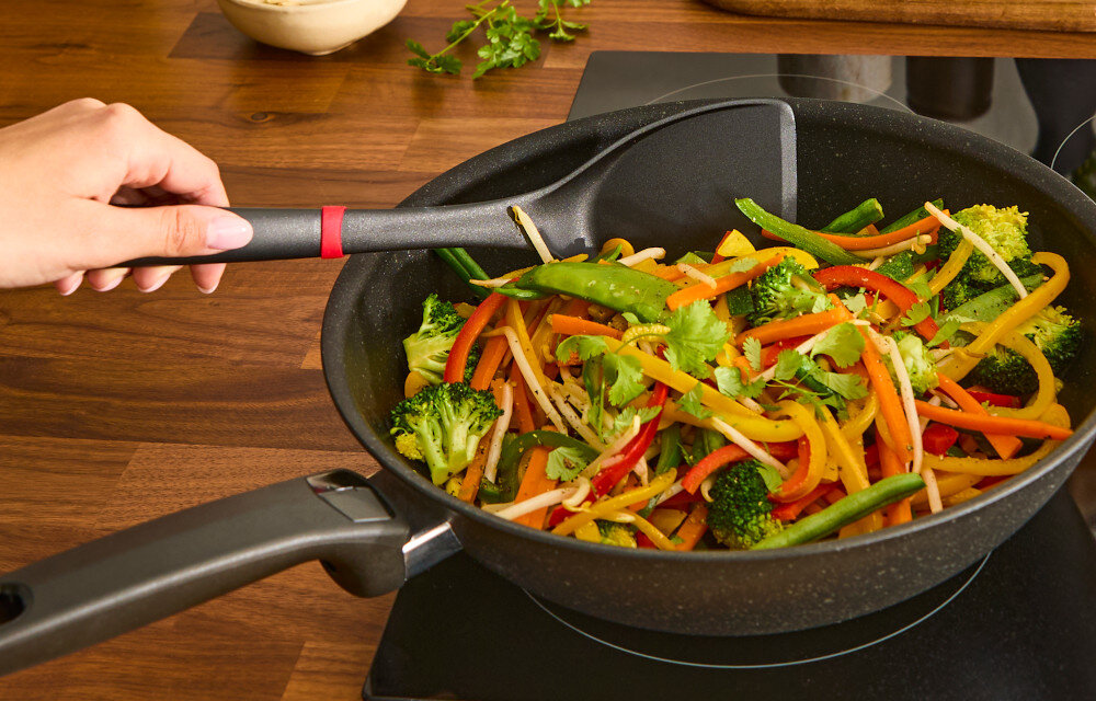 Osoba trzyma patelnię z kolorowymi warzywami, takimi jak papryka, brokuły i groszek cukrowy, na kuchence indukcyjnej. W tle widać drewniany blat z ziołami.