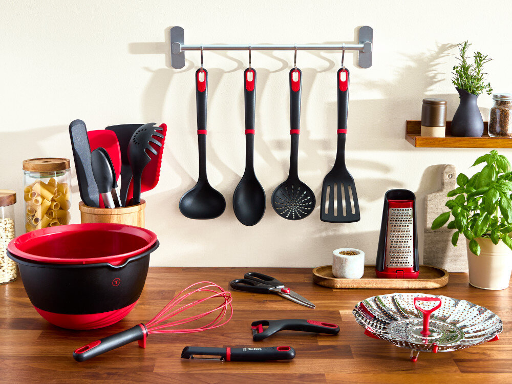 Na blacie kuchennym znajdują się różne przybory kuchenne, w tym miska, trzepaczka, tarka i narzędzia do gotowania, a na ścianie wisi zestaw łyżek i łopatek. Obok widoczna jest roślina doniczkowa i pojemniki z przyprawami.