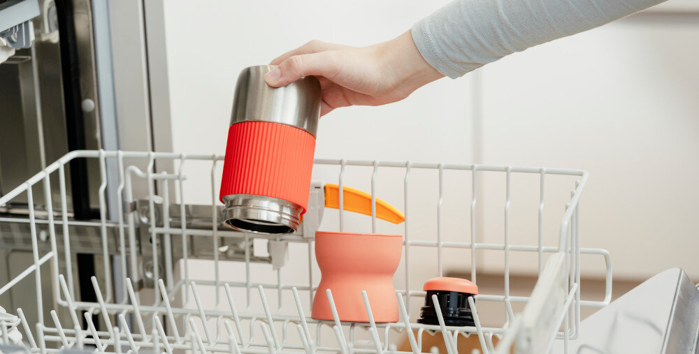 Osoba wkłada kubek termiczny z czerwonym uchwytem do zmywarki. W tle widoczne są inne elementy kuchenne.