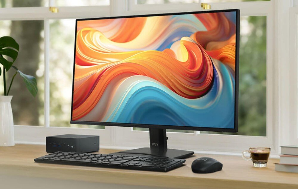 Monitor MSI Pro MP272 E14C Zdjęcie monitora na drewnianym blacie przy oknie, obok którego stoi miniaturowy komputer, czarna klawiatura, myszka oraz filiżanka kawy, opis