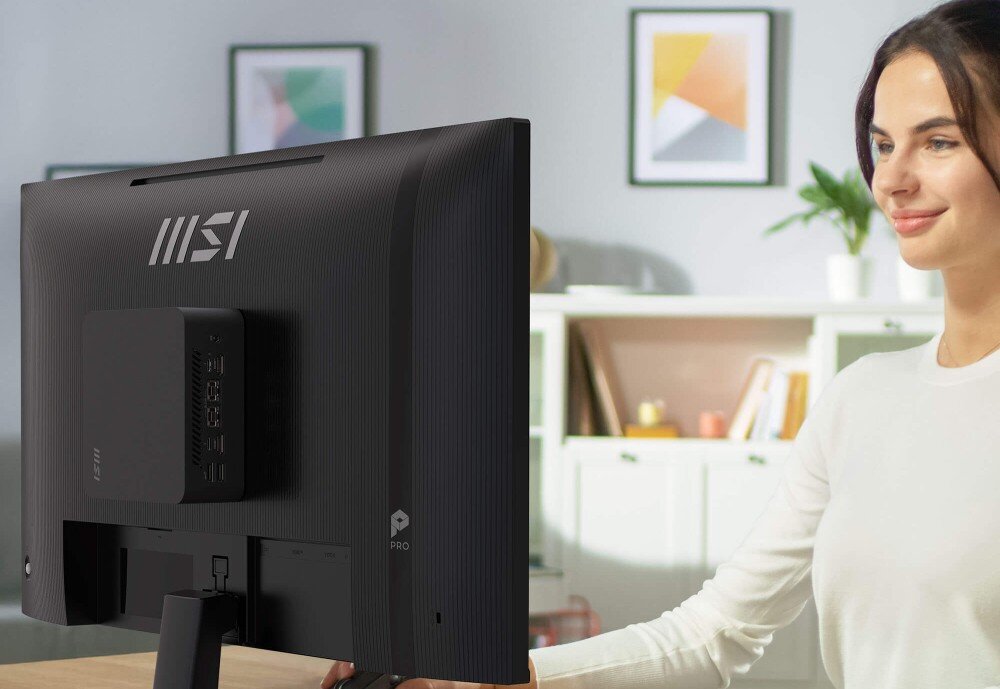 Monitor MSI Pro MP272 E14C Widok od tyłu na czarny monitor z zamontowanym na obudowie małym komputerem typu mini-PC, obok którego siedzi uśmiechnięta kobieta, montaż komputera, kompatybilność
