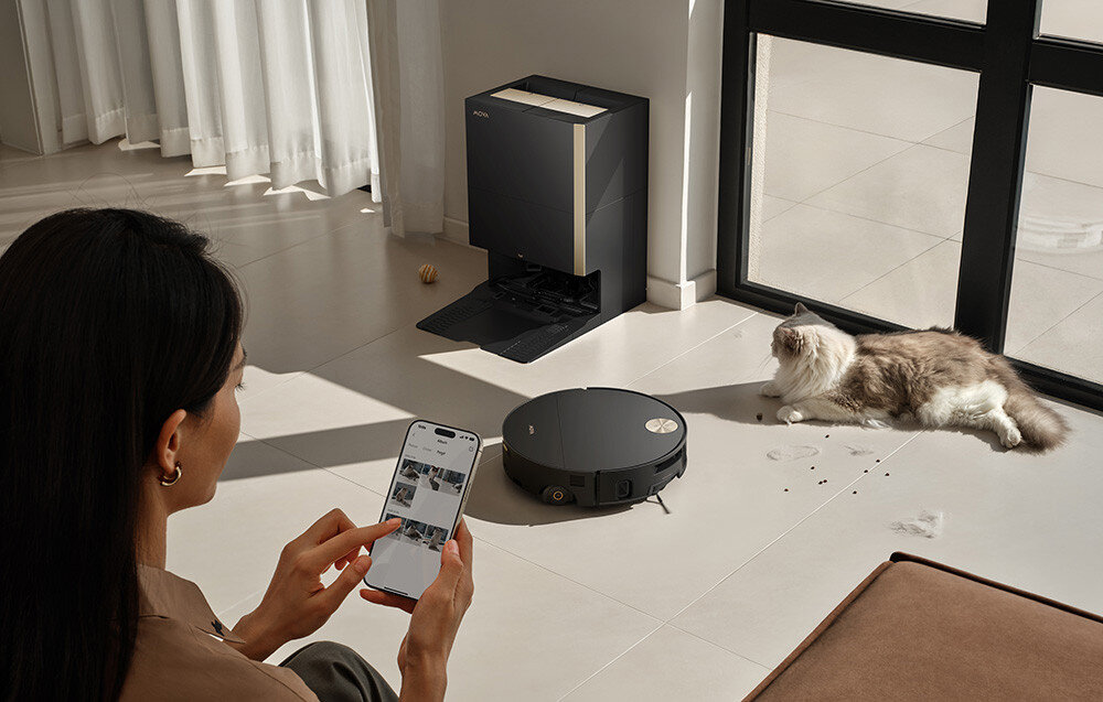 Na podłodze w jasnym pomieszczeniu znajduje się czarny robot sprzątający obok leżącego kota. W tle widać stację dokującą, a osoba trzyma w ręku smartfon z widocznymi obrazami na ekranie.