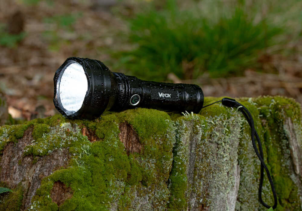 Czarna latarka leży na porośniętym mchem pniu drzewa. Na latarce widoczne jest logo \'Vayox\'.