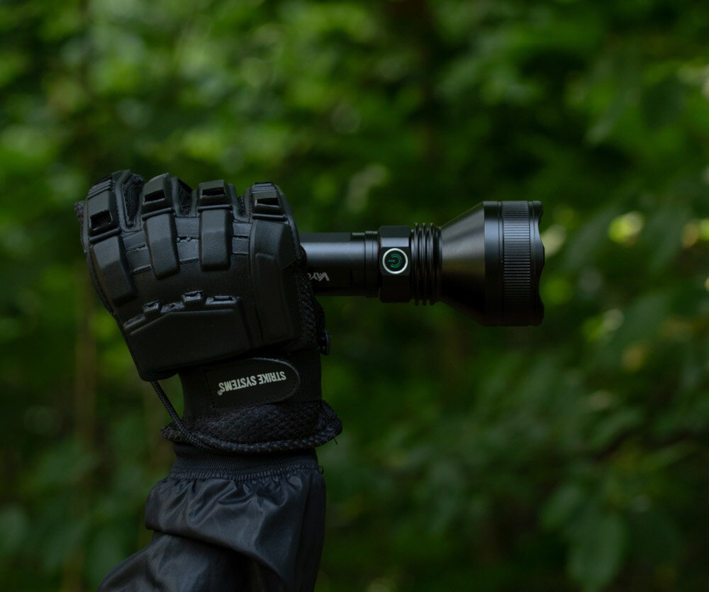 Osoba w czarnej rękawicy trzyma czarną latarkę z widocznym przyciskiem z zielonym symbolem. W tle znajduje się rozmyte zielone liście.