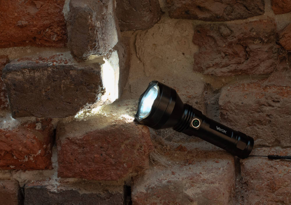 Czarna latarka leży na ceglanej ścianie, emitując jasne światło. Na latarce widoczne jest logo \'Vayox\'.