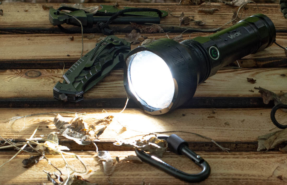 Na drewnianej powierzchni leży włączona latarka z widocznym logo \'VAYOX\', obok której znajdują się składany nóż i karabińczyk. Wokół rozrzucone są suche liście.
