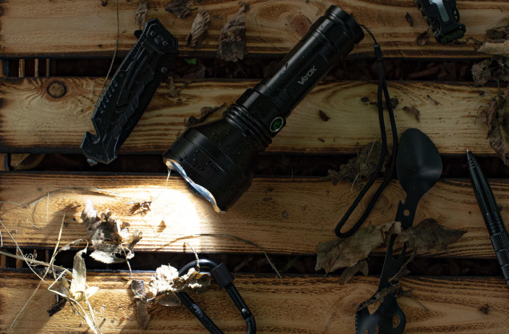 Na drewnianej powierzchni leży czarna latarka z napisem \'VAYOX\', obok której znajdują się różne narzędzia i liście. Latarka emituje światło, a wokół niej widoczne są suche liście.