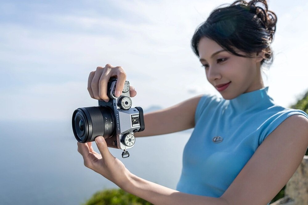 Osoba trzyma aparat fotograficzny z obiektywem, na tle rozmytego krajobrazu. Aparat ma widoczne pokrętła i wyświetlacz na górnej części obudowy.