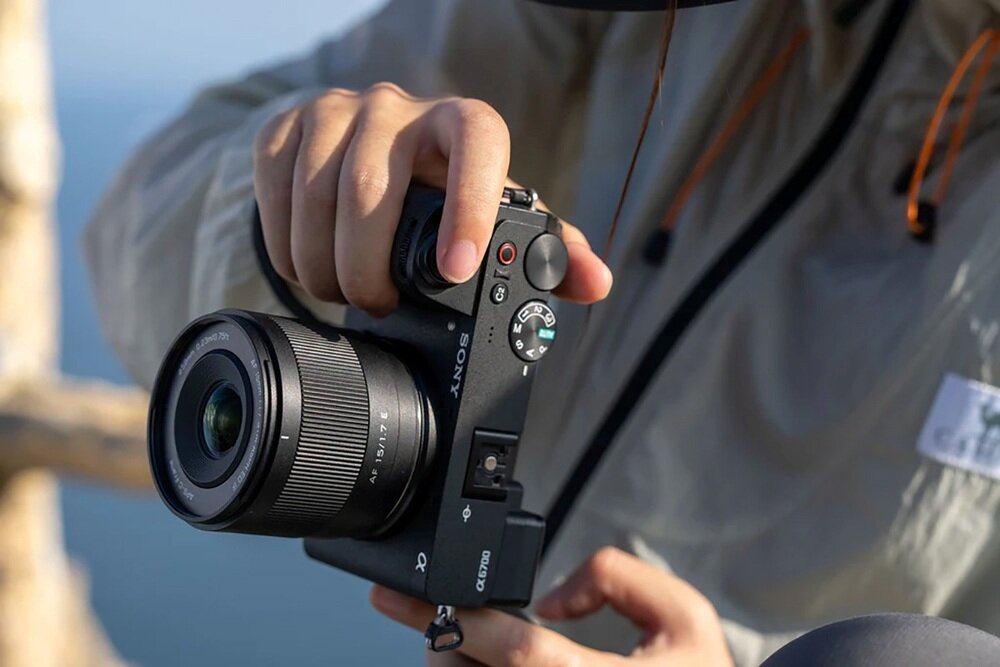 Osoba trzyma aparat fotograficzny z obiektywem, na którym widoczny jest napis \'AF 15/1.7\'. Na korpusie aparatu widoczne jest logo \'SONY\'.