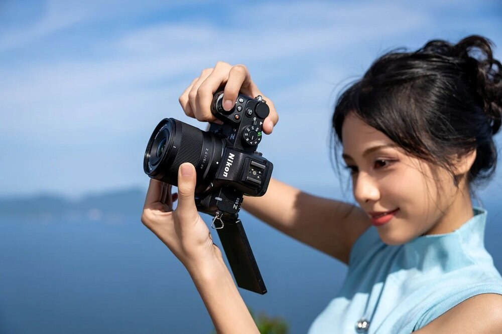 Osoba trzyma aparat fotograficzny z widocznym logo \'NIKON\' na tle nieba i wody. Aparat jest skierowany w stronę osoby, która patrzy na ekran.