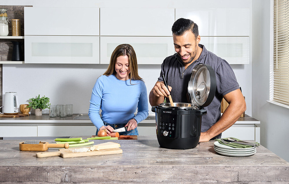 Dwie osoby przygotowują posiłek w kuchni, używając czarnego multicookera marki Instant Pot. Na blacie znajdują się deski do krojenia, warzywa i talerze.