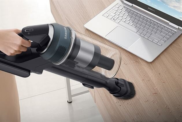 Osoba używa odkurzacza ręcznego marki Samsung do czyszczenia blatu stołu obok laptopa. Na blacie widoczne są drobne okruchy, które są zasysane przez odkurzacz.