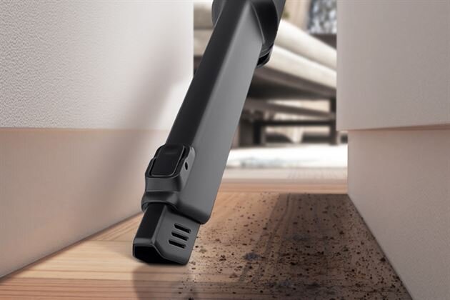 Czarna końcówka odkurzacza wąska, przystosowana do czyszczenia trudno dostępnych miejsc, znajduje się pomiędzy dwoma powierzchniami. Na podłodze widoczne są drobne zanieczyszczenia.