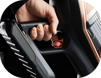 Osoba trzyma uchwyt odkurzacza z widocznym przyciskiem w kolorze czerwonym. Obudowa odkurzacza jest w kolorze czarnym z miedzianymi elementami.