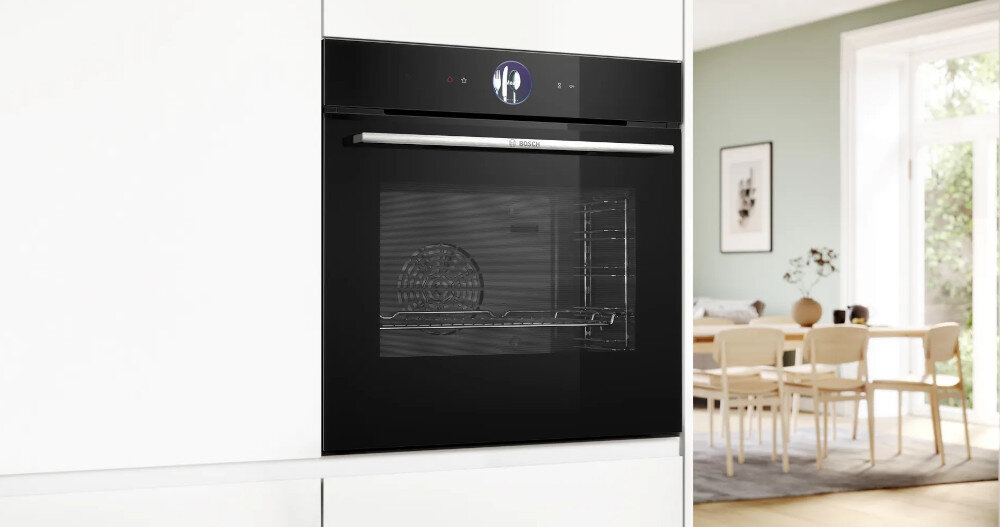 Czarny piekarnik elektryczny w zabudowie z widocznym logo BOSCH na uchwycie. W tle jasne wnętrze z dużym oknem i stołem z krzesłami.