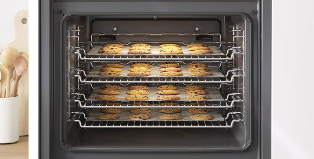 Wnętrze piekarnika z czterema poziomami rusztów, na których znajdują się blachy z piekącymi się ciasteczkami. Obok piekarnika widoczny jest stojak z drewnianymi łyżkami i deskami do krojenia.