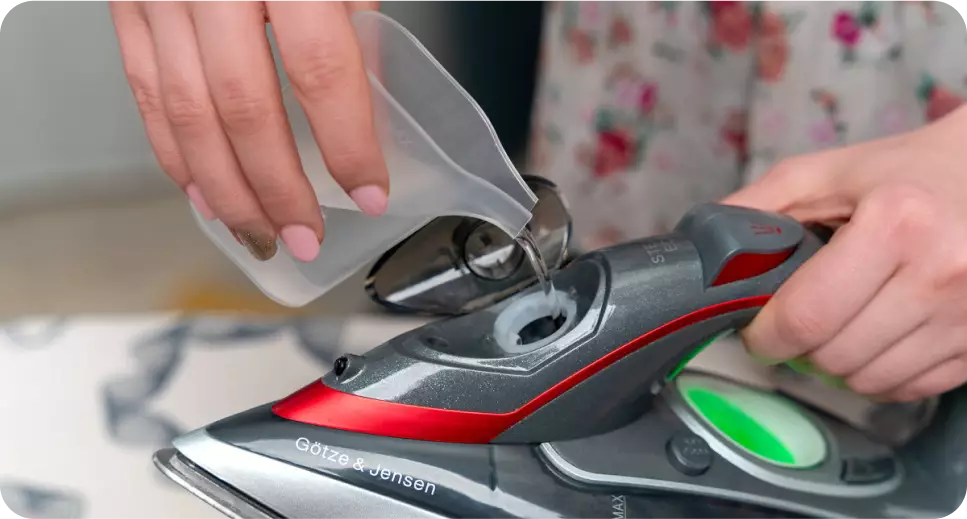Osoba nalewa wodę do żelazka marki Götze & Jensen. Na żelazku widoczny jest napis \'Götze & Jensen\'.