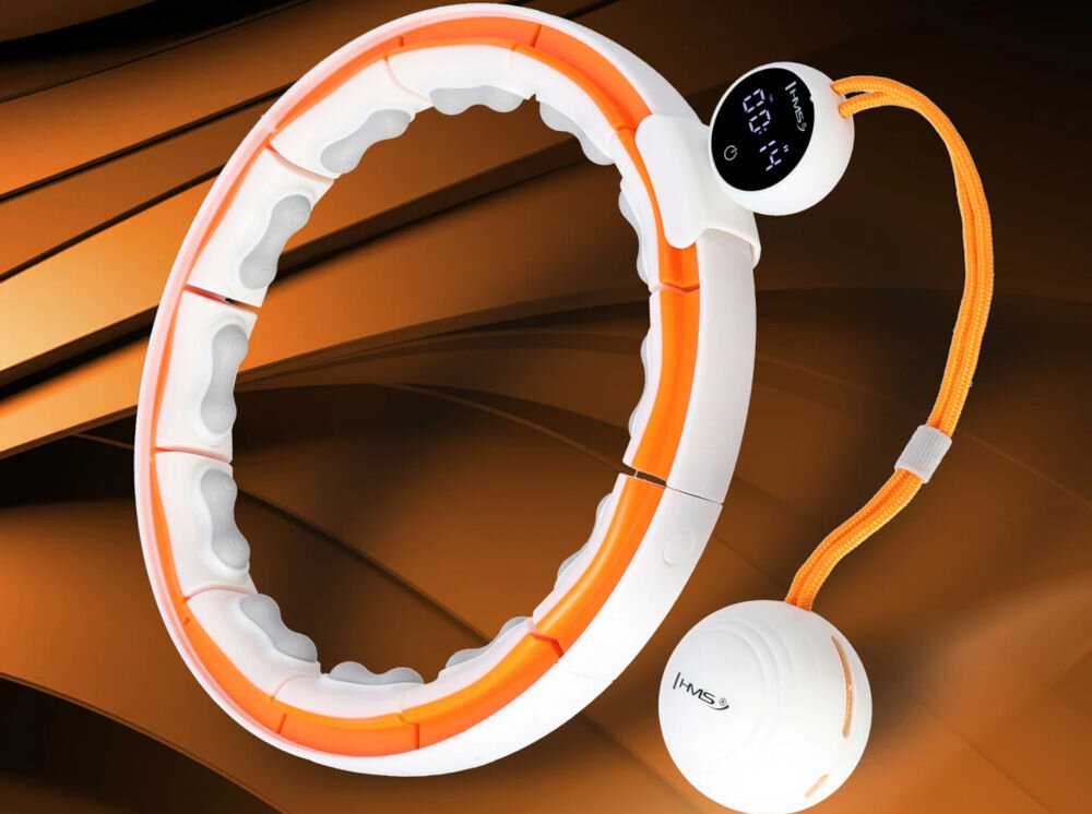 Na obrazie znajduje się biało-pomarańczowe hula hop z wyświetlaczem cyfrowym i przymocowaną kulką na sznurku. Na kulce widoczne jest logo \'HMS\'.