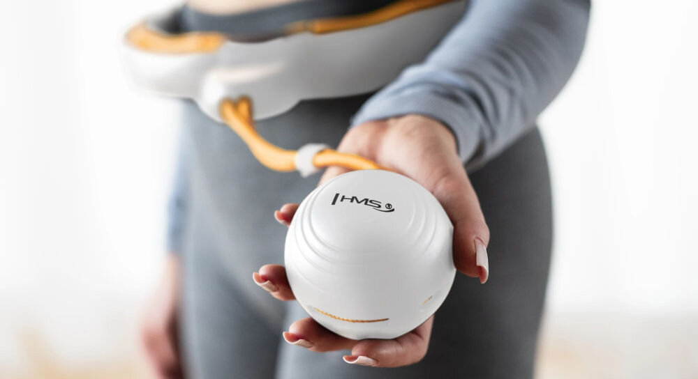 Osoba trzyma biało-pomarańczowy element hula hop z logo \'HMS\'. W tle widoczny jest pas hula hop wokół talii.