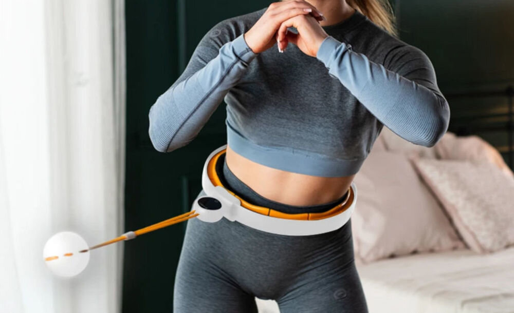 Osoba ćwiczy z hula hop w kolorze biało-pomarańczowym, które jest założone na talii. W tle widoczne są jasne zasłony i łóżko z poduszkami.