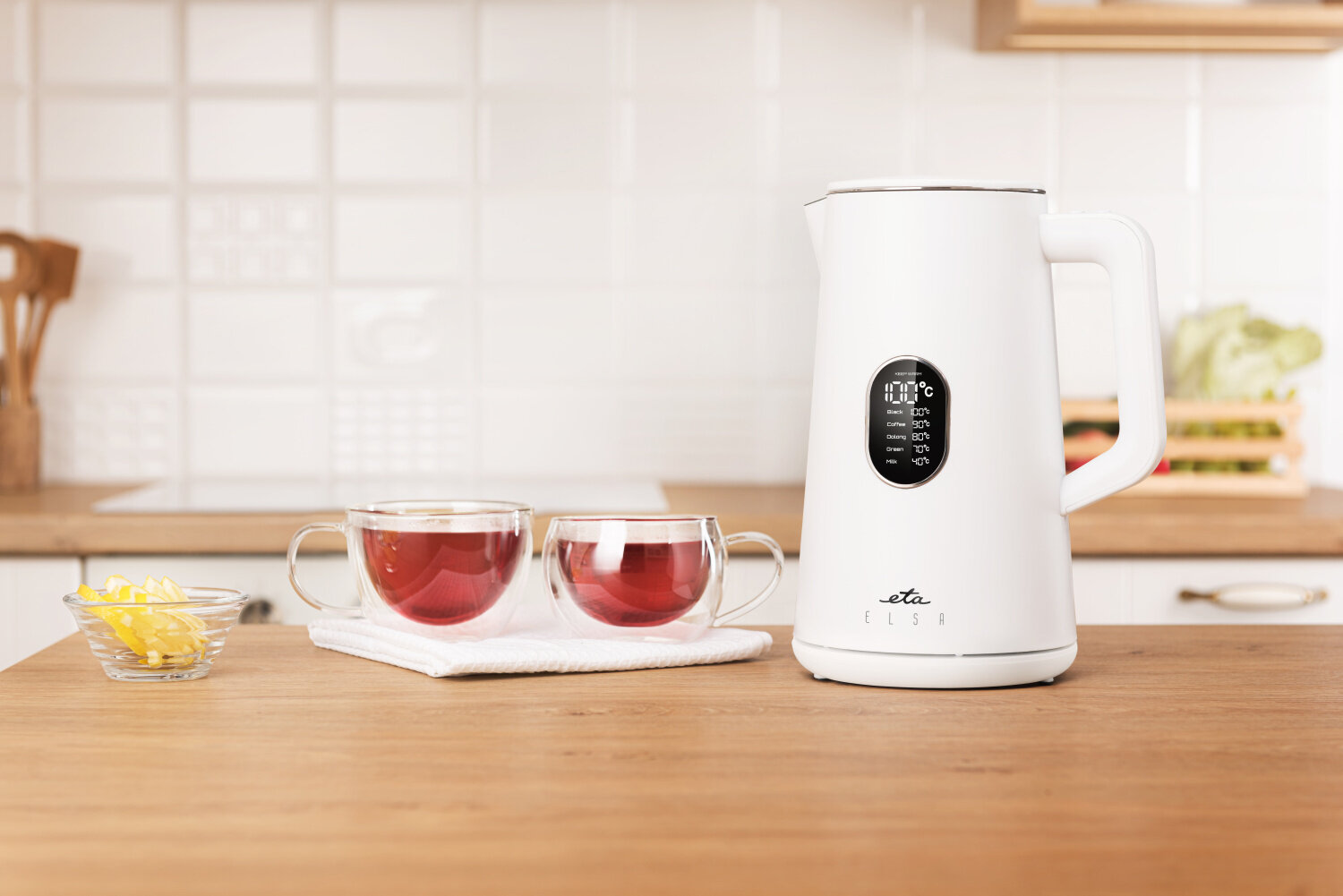 Na drewnianym blacie stoi biały czajnik elektryczny z wyświetlaczem oraz logo \'eta elsa\'. Obok znajdują się dwie szklanki z herbatą i mała miseczka z plasterkami cytryny.