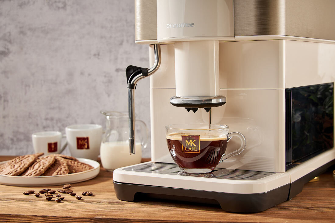 Ekspres do kawy w kolorze białym nalewa kawę do szklanej filiżanki z logo \'MK Cafe\'. W tle widoczne są filiżanki, dzbanek z mlekiem i talerz z ciastkami.