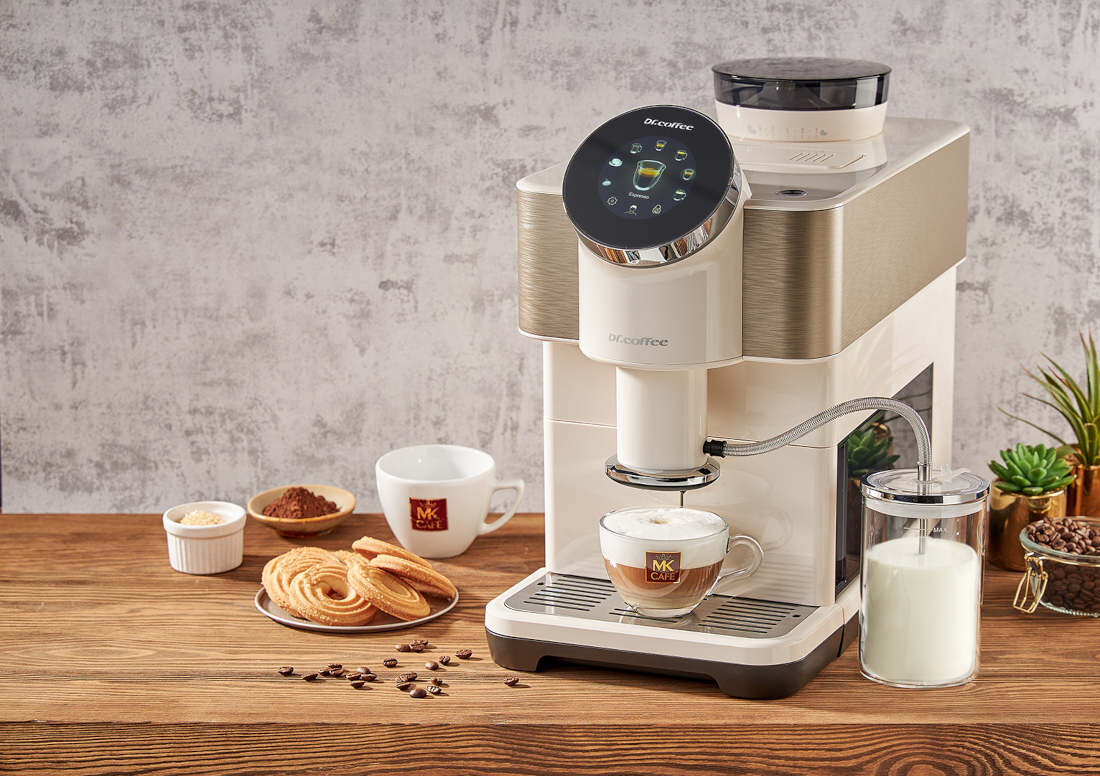 Na drewnianym blacie stoi ekspres do kawy DR.COFFEE H2 Biały z filiżanką kawy pod dyszą. Obok znajdują się filiżanki z logo MK Cafe, talerz z ciastkami, pojemnik z mlekiem oraz miseczki z kawą i cukrem.