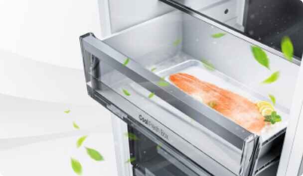 Otwarta szuflada lodówki z przezroczystym frontem, w której znajduje się kawałek łososia z plasterkiem cytryny. Wokół unoszą się zielone liście, a na szufladzie widoczny jest napis \'Cool Fresh Box\'.