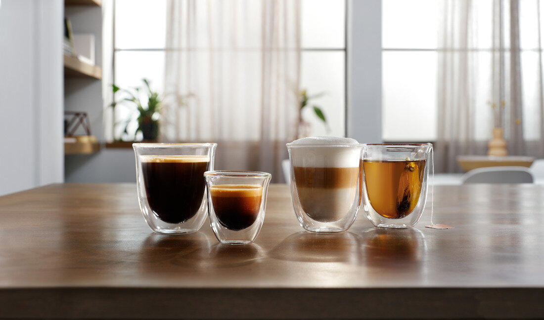 Na drewnianym stole stoją cztery szklanki z różnymi napojami. W tle widać zasłony i rozmyte elementy wnętrza.