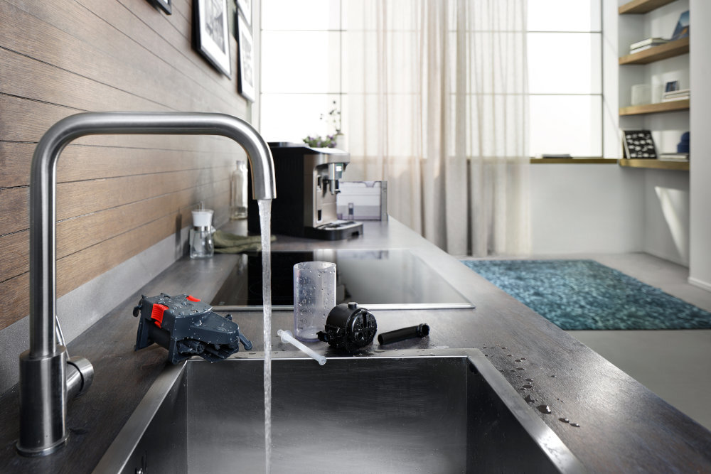 Kuchnia z widocznym zlewem, z którego leci woda, oraz ekspresem do kawy na blacie. Na zlewie znajdują się części do czyszczenia ekspresu.