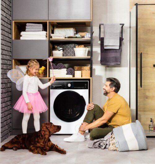 Pralka w zabudowie z czarnym panelem sterowania i dużym okrągłym bębnem. Obok pralki znajdują się półki z ręcznikami i koszami, a na podłodze siedzi mężczyzna z dzieckiem i psem.