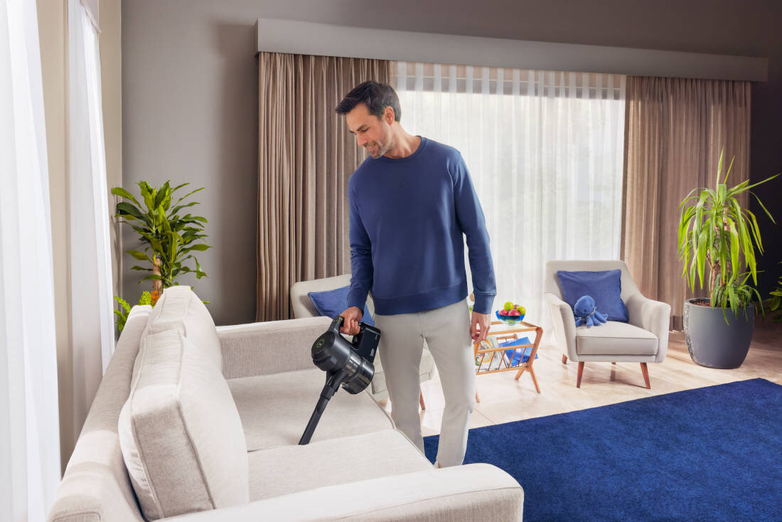 Osoba używa odkurzacza ręcznego do czyszczenia jasnej kanapy w salonie. W tle widać zasłony, rośliny doniczkowe i dwa fotele z poduszkami.