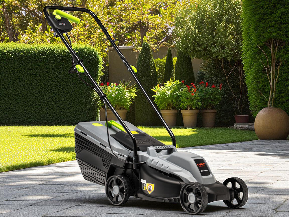 Elektryczna kosiarka z czarnym i szarym korpusem oraz zielonymi akcentami stoi na brukowanej powierzchni w ogrodzie. W tle widoczne są zielone krzewy i kwiaty w donicach.