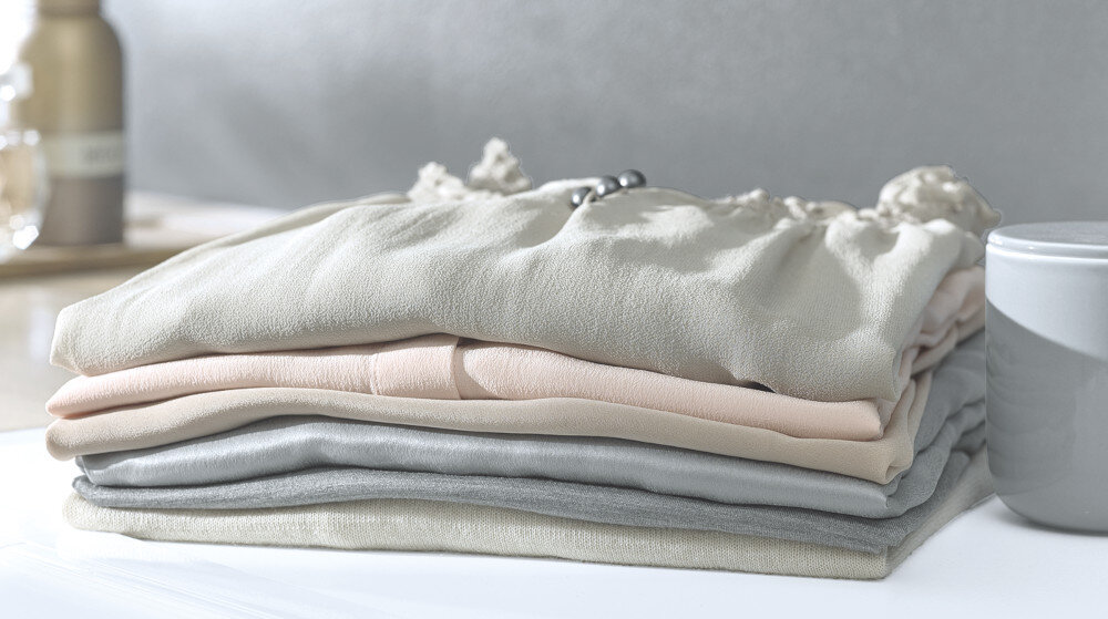 Na zdjęciu widoczny jest stos złożonych ręczników w różnych odcieniach beżu i szarości. Obok znajduje się szara, cylindryczna pojemnik.