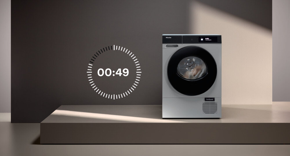 Na zdjęciu znajduje się suszarka z widocznym logo \'Miele\' oraz napisem \'EcoSpeed\'. Obok suszarki widoczny jest zegar z czasem \'00:49\'.