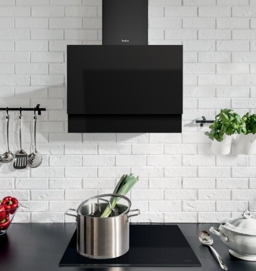 Czarny okap kuchenny marki Amica zamontowany na białej ceglanej ścianie nad płytą indukcyjną. Na płycie stoi garnek z warzywami, a obok znajdują się przybory kuchenne i doniczka z ziołami.