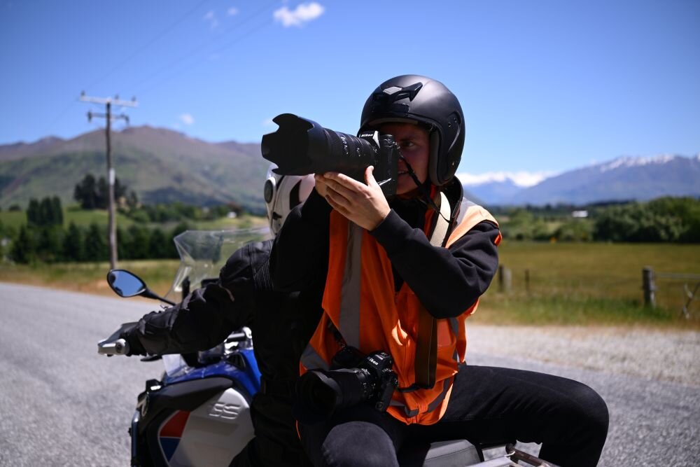 Osoba w kasku i pomarańczowej kamizelce siedzi na motocyklu i trzyma aparat z dużym obiektywem. W tle widać zielone pola i góry pod błękitnym niebem.