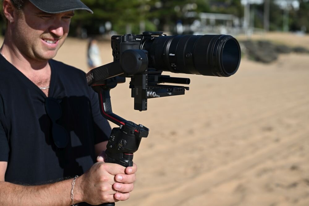 Osoba trzyma aparat z dużym obiektywem zamontowanym na stabilizatorze, na tle piaszczystej plaży. Na obiektywie widoczne jest logo \'Nikon\'.