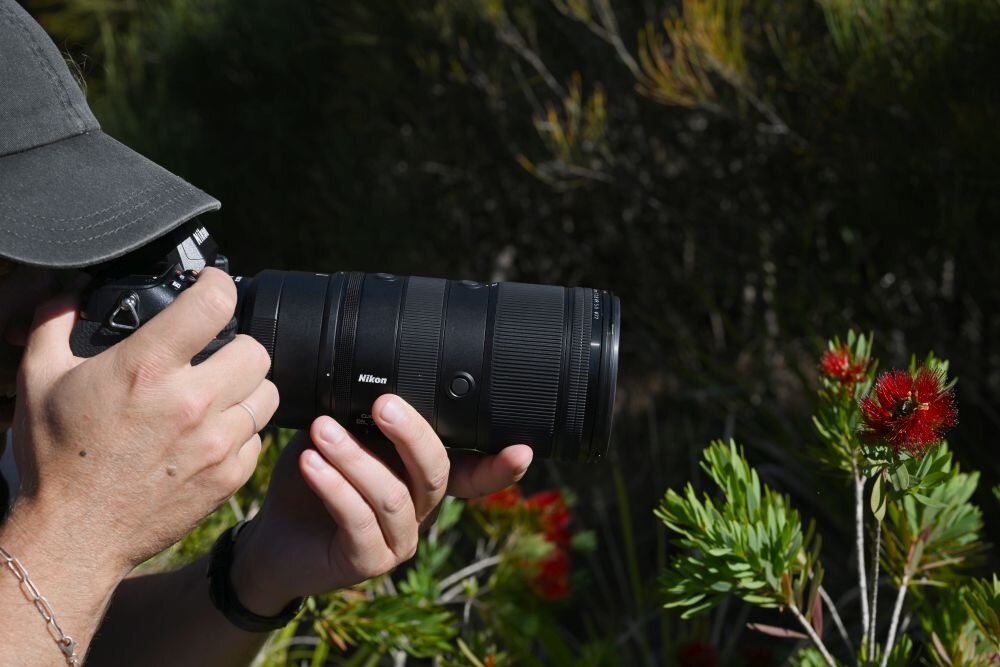 Osoba trzyma aparat fotograficzny z dużym obiektywem marki Nikon, skierowanym w stronę roślin z czerwonymi kwiatami. W tle widoczne są zielone krzewy.