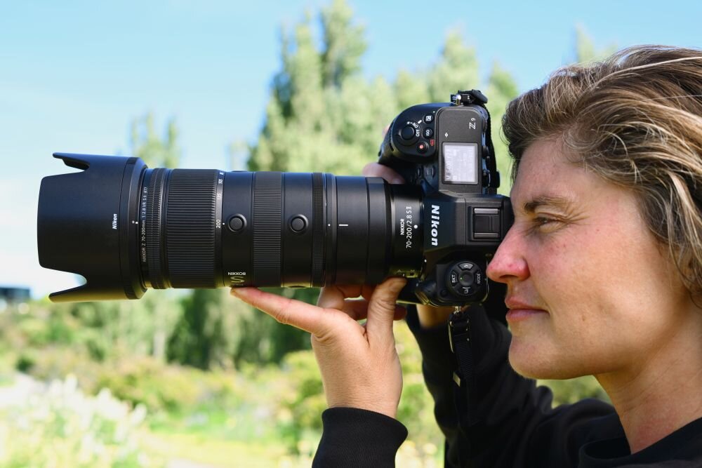 Osoba trzyma aparat fotograficzny z dużym obiektywem marki Nikon, na tle zielonego krajobrazu. Na obiektywie widoczne są napisy: \'NIKKOR Z 70-200mm f/2.8 VR S\'.