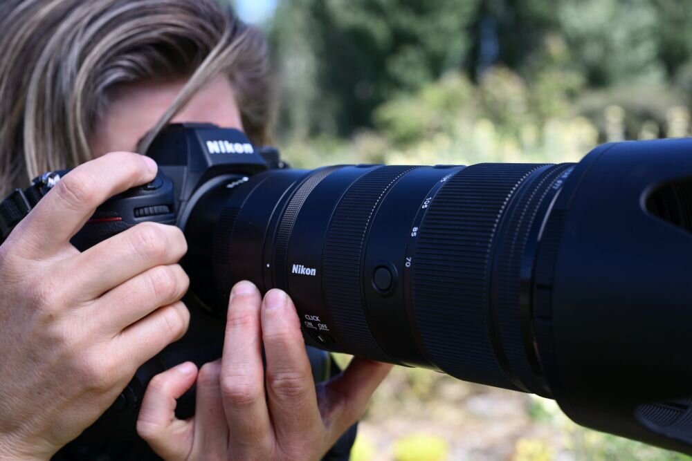 Osoba trzyma aparat fotograficzny z dużym obiektywem marki Nikon. W tle widoczna jest rozmyta zieleń.