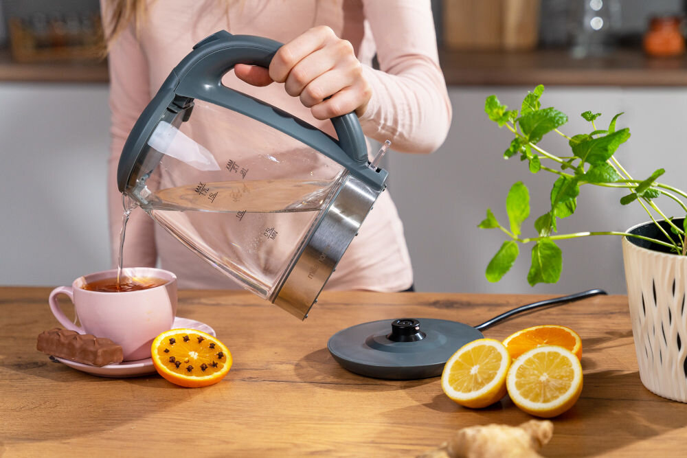 Osoba nalewa wodę z czajnika do różowej filiżanki z herbatą, obok której leży plasterek pomarańczy i baton. Na stole znajdują się również plasterki cytryny i pomarańczy oraz doniczka z rośliną.