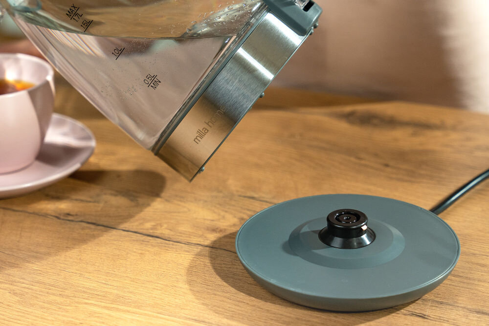 Szklany czajnik z miarką i napisem \'milla home\' na drewnianym blacie obok podstawki. W tle różowa filiżanka na spodku.