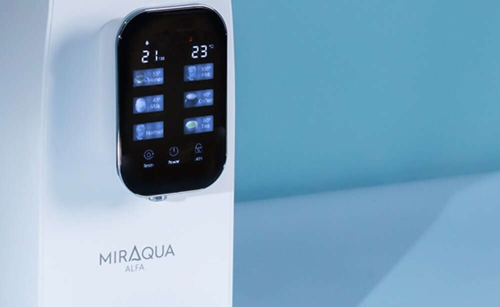 Biały dyspenser do wody z wyświetlaczem dotykowym pokazującym temperatury 21 i 23 stopnie oraz różne opcje ustawień. Na obudowie widoczny jest napis \'MIRAQUA ALFA\'.
