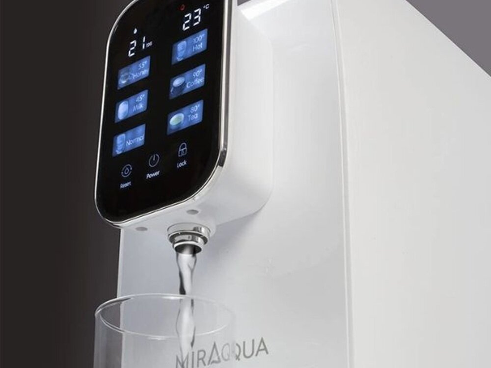 Na obrazie widoczny jest dyspenser do wody z wyświetlaczem dotykowym, z którego nalewana jest woda do szklanki. Na urządzeniu widnieje napis \'MIRAQUA\'.