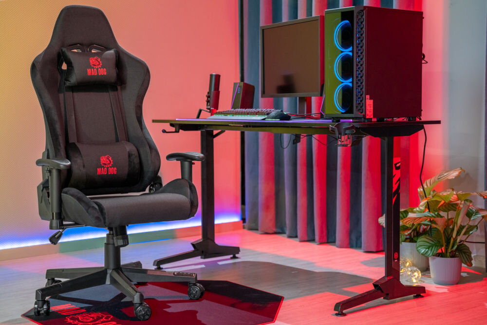 Na obrazie znajduje się czarny fotel gamingowy z logo \'MAD DOG\' na zagłówku i oparciu. Obok fotela stoi biurko z komputerem, monitorem i klawiaturą, a w tle widoczne są zasłony i rośliny.