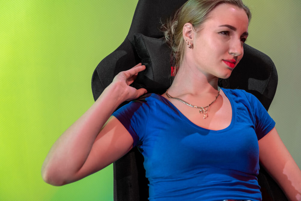 Osoba siedzi na czarnym fotelu gamingowym z wysokim oparciem. Tło jest w kolorze zielonym.