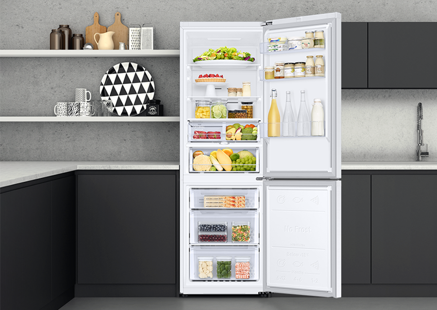 Lodówka z wygodnym dostępem do produktów Biała lodówka z otwartymi drzwiami, wypełniona różnorodnymi produktami spożywczymi, takimi jak owoce, warzywa, napoje i słoiki. W tle widoczne są szare ściany i czarne szafki kuchenne.