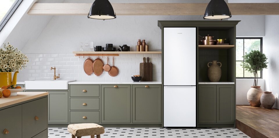 Lodówka wolnostojąca Samsung RB34C603CWW/EF dostępna w sklepach Media Expert Kuchnia z zielonymi szafkami i białą lodówką w zabudowie. Na blacie stoją naczynia i dekoracje, a na ścianie wisi półka z dodatkami kuchennymi.